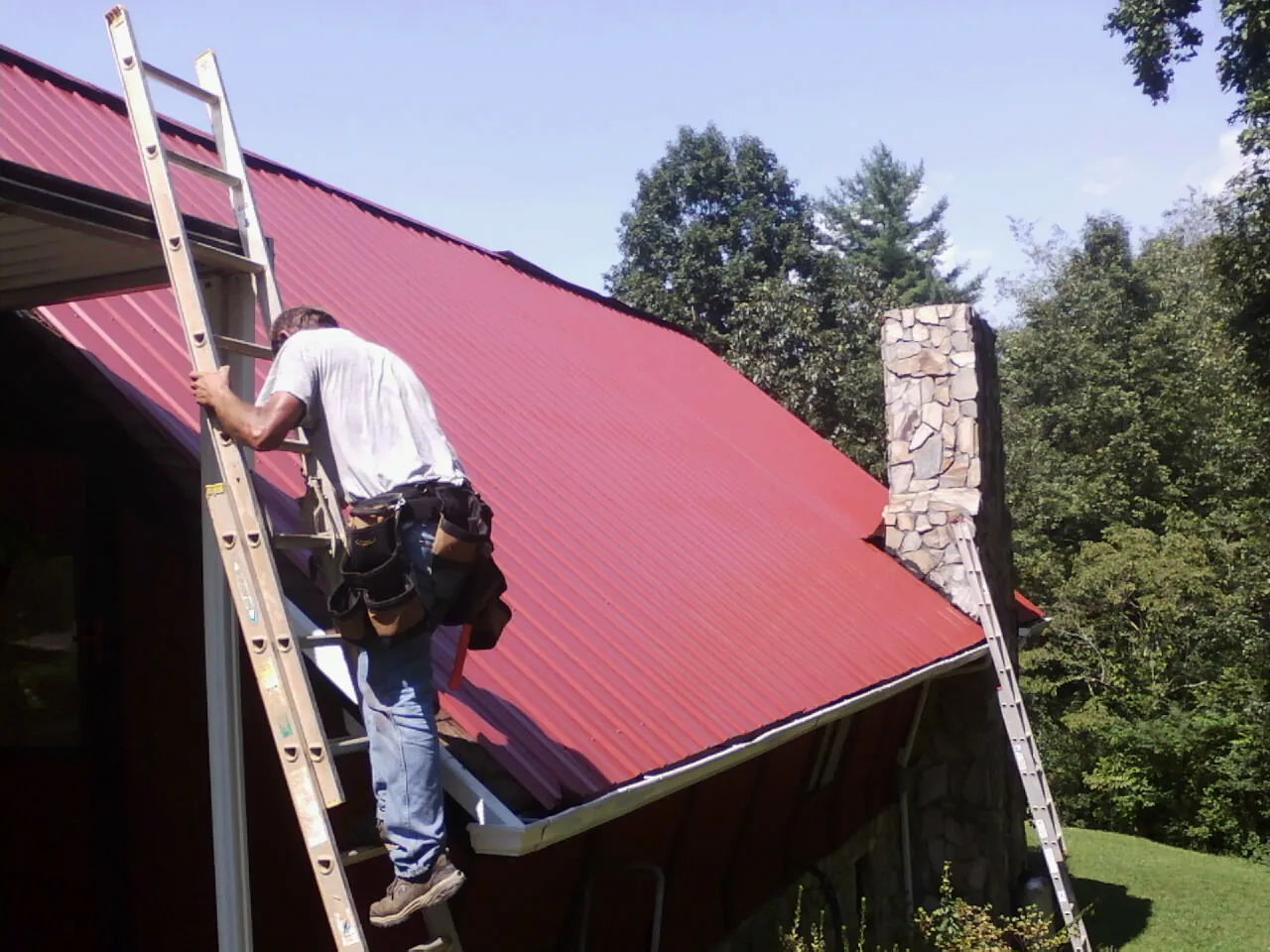 Roofer on a ladder beside a red metal roof and stone chimney — Soffit & Fascia Repair by Roofing Brothers in Ironside